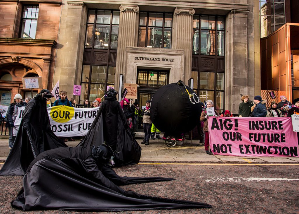 Protest outside insurance company AIG office. Activists hold placards and banners saying AIG! Insure our Future not Extinction and Insure our Future not Fossil Fuels. Oil Slicks, a protest troupe dressed head to toe in black, perform in front of protest with one kneeling on road. There is a large carbon bomb prop saying CO2 on it.